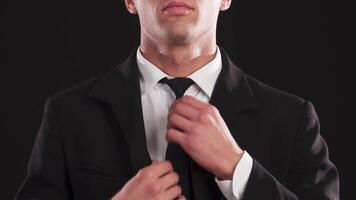 A dapper young man adjusts his tie with precision, exuding charm and sophistication against a sleek black background. His half-visible face radiates confidence, complemented by a broad smile that enhances his handsome features video