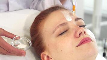 A skilled cosmetologist in a crisp white coat gently applies a creamy facial mask to the forehead of a serene young woman. This close-up captures the essence of beauty and relaxation, showcasing the delicate process of skin rejuvenation and hydration video