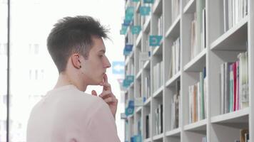 Thoughtful young man exploring bookshelves in a bright library video