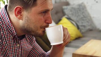 The young man with a stylish checkered shirt sits comfortably in a cozy cafe, relishing every sip of his aromatic coffee. His expression radiates satisfaction as he enjoys the rich flavors, surrounded by a warm and inviting atmosphere video