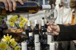Close-up of a person's hand pouring white wine into a glass, many bottles of wine nearby. Yellow flowers in the background, shallow depth of field. The image conveys the elegance of a fine dining experience. photo