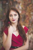 portrait of a young girl in a red dress against the background of autumn trees with a rose in her hands. thoughtful and romantic girl with long dark hair. emphasis on the red dress, lips, rose photo