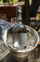 A bottle of champagne stands in an ice bucket, outdoors. The setting suggests a bright celebration, with champagne glasses in the background. photo