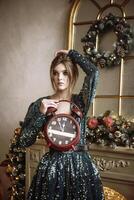 young brunette in a green evening dress with a big round watch in her hands, showing five to twelve. she is standing against the background of a fireplace and New Year's decorations.she is making a wish photo
