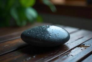 A black stone with water drops on a wooden surface, in a blurred background with green leaves photo