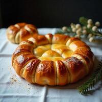 A ring-shaped bread with a golden crust, placed on a white tablecloth, with a dark background and a sprig of green leaves photo