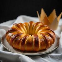 A ring-shaped sweet bread with a golden crown in the background, on a white tablecloth with some green branches photo