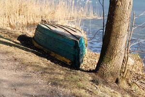 Vintage green boat with yellow stripe stored upside down under tree on riverbank near dirt path. Early spring scene. Design for postcard, greeting card Side view composition photo