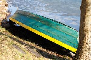 Old green and yellow boat turned upside down on lakeside grass near tree trunk. Early spring outdoor photography. Rustic fishing concept. Design for postcard, wallpaper, greeting card, banner photo