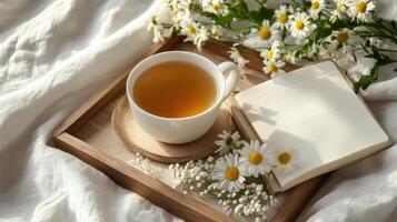 Cozy scene featuring cup of chamomile tea on wooden tray, surrounded by fresh chamomile flowers and open notebook photo