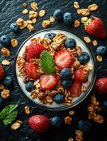 cuenco de cremoso yogur coronado con Fresco fresas, arándanos, Granola, y menta hoja, metido en oscuro texturizado superficie foto