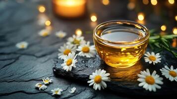 Glass bowl of golden essential oil surrounded by chamomile flowers on dark stone surface photo