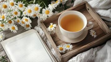 Cozy scene with cup of chamomile tea on wooden tray, surrounded by fresh chamomile flowers, notebook, and soft fabric photo