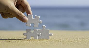 Hand placing a white jigsaw puzzle piece on top of two others on sand, completing a trio. Blurred blue sea background. Concept of problem solving and teamwork. photo