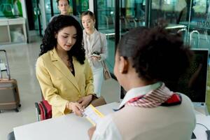 Female traveler checking in at airport counter, handing over boarding pass to staff, with passengers waiting in line and suitcases visible in modern terminal photo