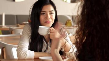 A cheerful Asian girl beams with laughter as she engages in a lively conversation with her curly-haired friend at a trendy cafe. The warm ambiance, with soft lighting and stylish decor, enhances the joyful atmosphere of friendship and connection video