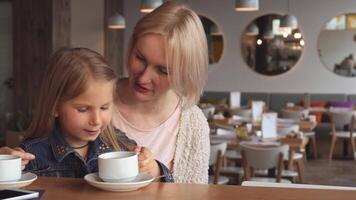 A charming little girl sips tea from a delicate white cup while her mother shares an engaging story. The warm atmosphere of the cafe, with soft lighting and inviting decor, enhances their joyful bonding moment video