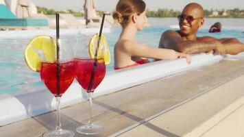 A vibrant multiethnic couple relaxes in a shimmering swimming pool, sharing laughter and cocktails. The scene is beautifully framed by refreshing drinks adorned with lemon slices, evoking a sense of summer bliss and romantic getaway video