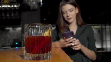 A stylish woman in a trendy outfit is poised at the bar, reaching for a vibrant cocktail glass as she texts on her smartphone. The scene captures the essence of modern nightlife, blending social interaction with the enjoyment of a refreshing drink video