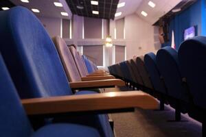 A conference room with rows of empty blue chairs and square lamps on the ceiling. With space to copy photo