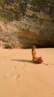 FPV drone shot of woman meditating alone on remote tropical beach with turquoise sea, waves, and cliffs, Bali, Indonesia. Peaceful scene of relaxation, mindfulness, and inner peace. video