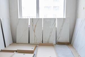 A large marble tile for walls measuring 120 by 60 is displayed in a row, the selection of the pattern on the tile for laying on the walls. On the background of a window under daylight photo
