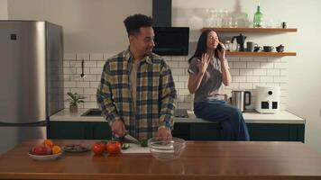 African American man prepares a fresh salad in the kitchen while smiling at his girlfriend, who sits on the counter talking on the phone. A joyful, sunlit morning scene full of warmth and connection. video