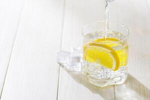 Pouring a glass of water with lemon and ice on a white table. photo