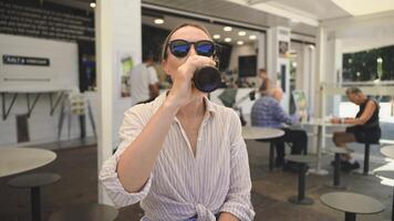 A young woman enjoys a refreshing drink from a bottle at a sunny outdoor caf , savoring her relaxation during a hot summer day. Perfect urban leisure moment with casual sophistication. video