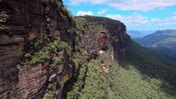 An aerial shot captures a dramatic cliff face towering above a lush green valley, with striking red rock formations, endless forests, and cloud-dotted skies. video