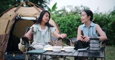 Handheld shot, Two Asian friends sit on chairs and talk happily in front of a tent in a camping atmosphere. video