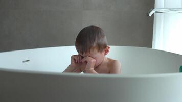 A little boy of 4 years old is happily bathing in a white oval bathtub, the water has gotten into his eyes and he is carefully wiping the water from his eyes with his fists video