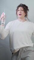 Woman in pajamas dances and sings joyfully while using an electronic water flosser for morning tooth care on gray background video