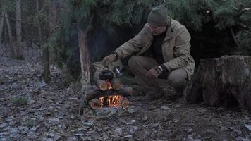 A man cooking food over a fire in the woods, squatting next to a shelter of pine branches, placing a kettle on burning logs in a forest clearing. video