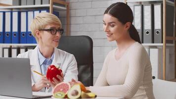 A dietitian consults with a client at a clinic, discussing healthy food choices. Fresh fruits and vegetables are present. The setting appears professional, emphasizing nutrition education. video