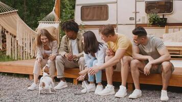 A group of diverse friends relax together at a camping site, engaging with a small dog, while enjoying the warmth of summer and the great outdoors. Laughter fills the air as they bond. video