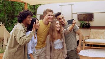A diverse group of friends is enjoying their time together outdoors at a campsite during the summer. They are laughing and posing for a selfie, capturing their joyful moments. video