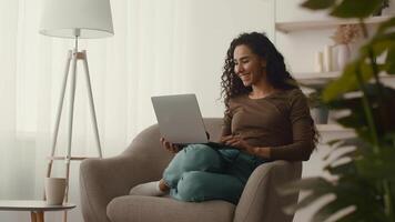 In a cozy living environment, a young woman sits comfortably in a plush chair, smiling as she interacts with her laptop. Sunlight pours in, highlighting her relaxed demeanor. video