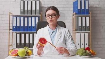 A dietitian in a white coat sits at a desk discussing the benefits of various fruits. Surrounding her are colorful fruits like apples and grapefruits, highlighting healthy eating. video