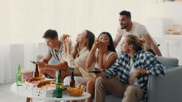 In a bright living room, a diverse group of friends share a relaxed evening filled with laughter, pizza, snacks, and drinks, all while engaging with their phones and each other. video
