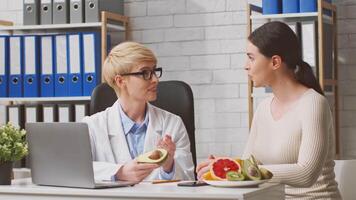 A dietitian consults with a woman patient in a bright clinic, discussing nutrition and healthy food choices. Fresh fruits and vegetables are on display, emphasizing a wholesome diet approach. video