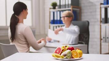 A healthcare professional guides a lady patient in a modern clinic, discussing dietary plans while colorful fruits are displayed on the table. The consultation emphasizes healthy food options. video