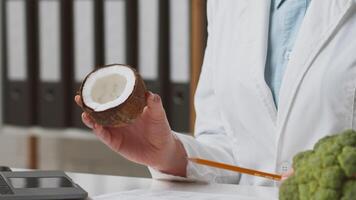 A healthcare professional in a white coat holds a coconut while explaining its nutritional benefits in a clinic. Broccoli and a laptop are visible on the table, showcasing healthy food choices. video