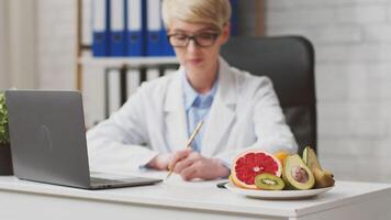 A healthcare professional, dressed in a white coat, is seated at a desk in a clinic, reviewing information on a laptop while taking notes. Fresh fruits are displayed nearby, emphasizing nutrition. video