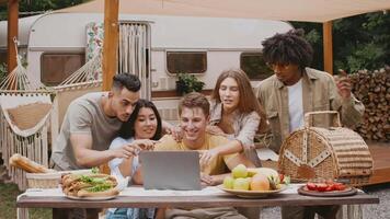 A diverse group of friends is gathered outdoors during a summer weekend, laughing and sharing experiences together while looking at a laptop and enjoying fresh fruit and snacks. video