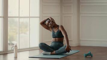 A young African American woman practices stretching exercises indoors in a bright space. She focuses on maintaining a healthy lifestyle while seated on a yoga mat. video
