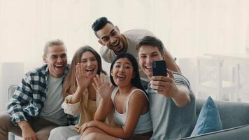 A group of diverse millennials is happily gathered together in a bright, modern living room, sharing laughs and taking a selfie. Their lively interaction reflects a joyful friendship. video