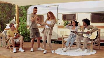 A diverse group of friends gathers on a wooden deck at a campsite, dancing and playing music while enjoying their weekend getaway in nature. Laughter fills the air as they relax together. video