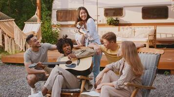A group of diverse friends relaxes in a camping setting, playing guitar and sharing moments of joy. The atmosphere is filled with laughter, with a cozy trailer in the background. video