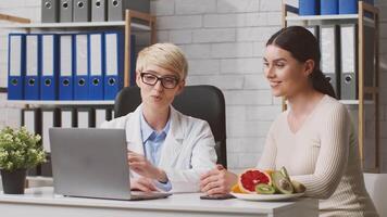 A dietitian in a white coat discusses dietary plans with a lady patient in a clinic. Fresh fruits and a computer are on the table, indicating a focus on health and nutrition. video